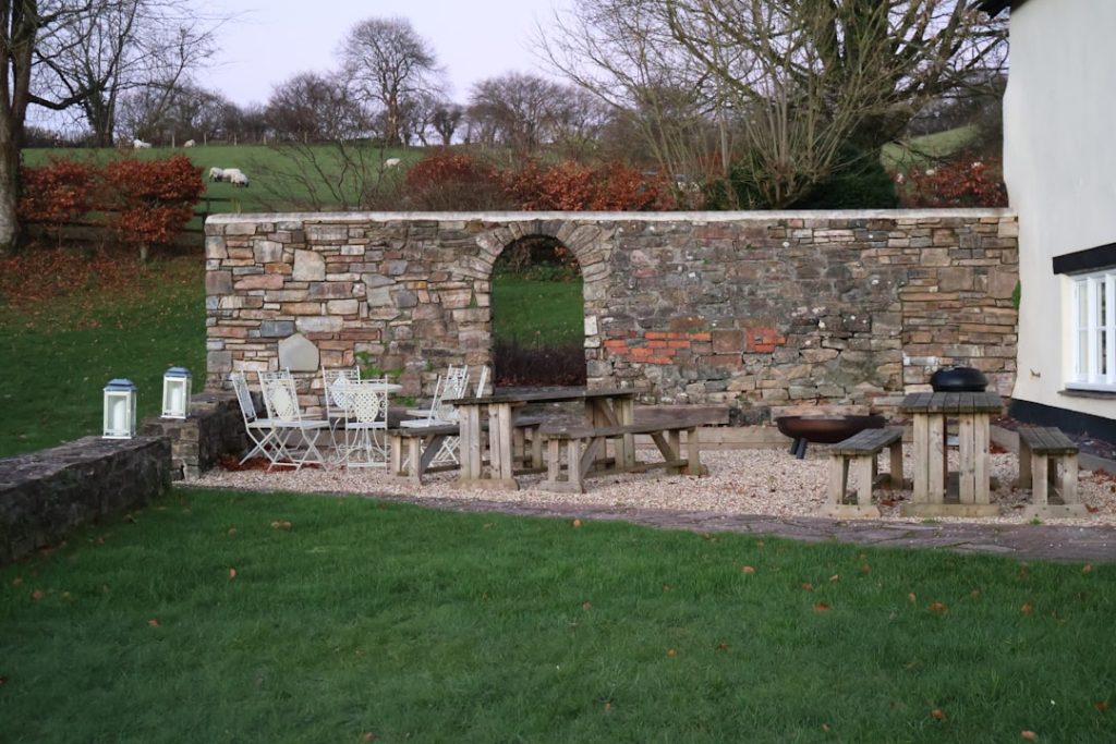 A cozy outdoor entertaining area featuring wooden picnic tables, chairs, and a stone wall backdrop in a grassy backyard.